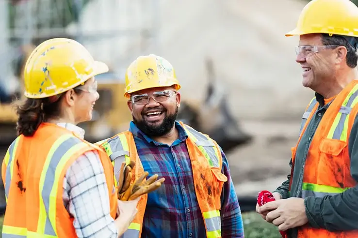 A group of three workers at a construction site wearing hard hats, safety glasses and reflective clothing, smiling and conversing.