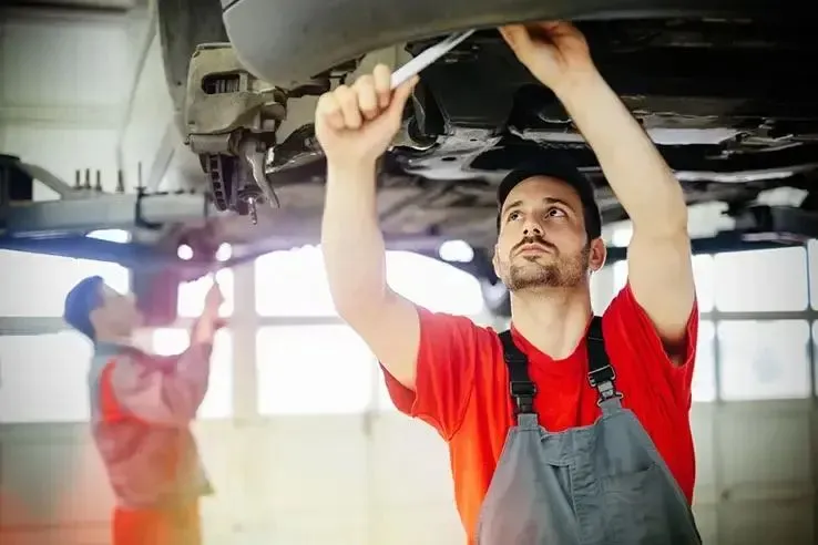 Two mechanics work on the underside of a car which has been lifted up above them in the garage.