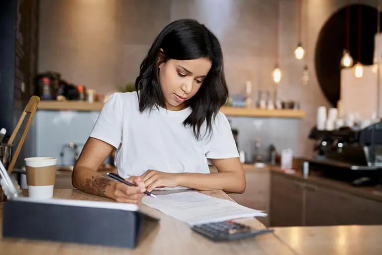 A woman sits in her pop-up shop with a coffee, working out her expenses with a calculator, and pen & paper.