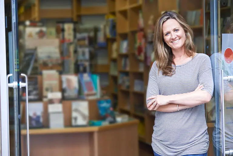 A woman stands smiling in the doorway to her pop-up shop which can be seen in the background, full of colourful bookshelf displays.