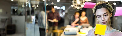 close-up of female employee posting sticky notes to a transparent board in a meeting room, surrounded by her colleagues in the background.
