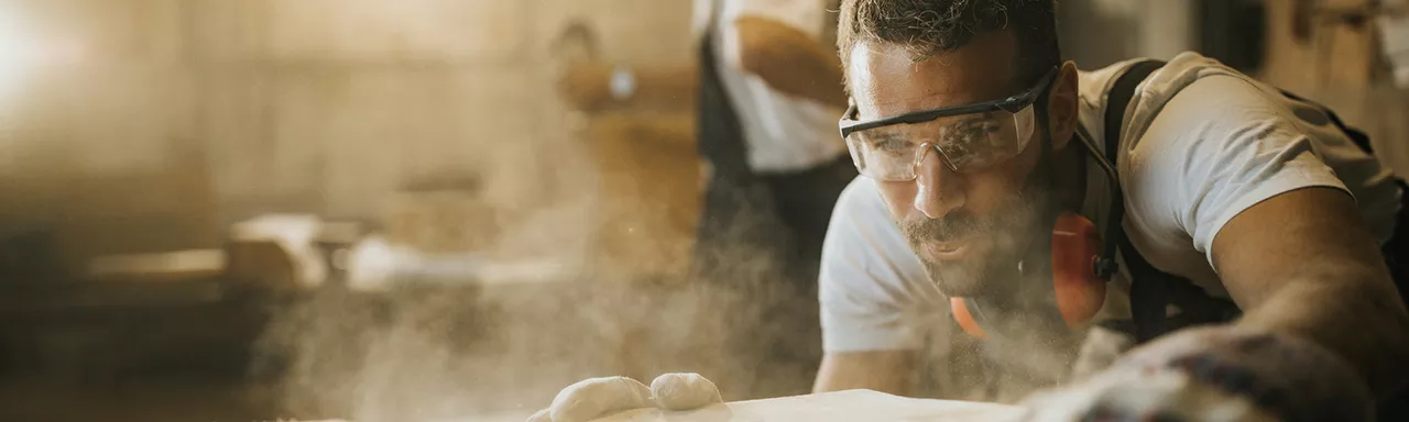 closeup of young carpenter wearing safety goggles sanding wood in his workshop with another worker in the background