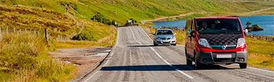 A red van is driving past a lake on a narrow country road.