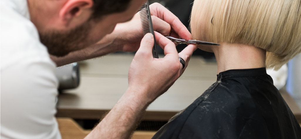 A hairdresser trimming the back of a person's blonde bob haircut.